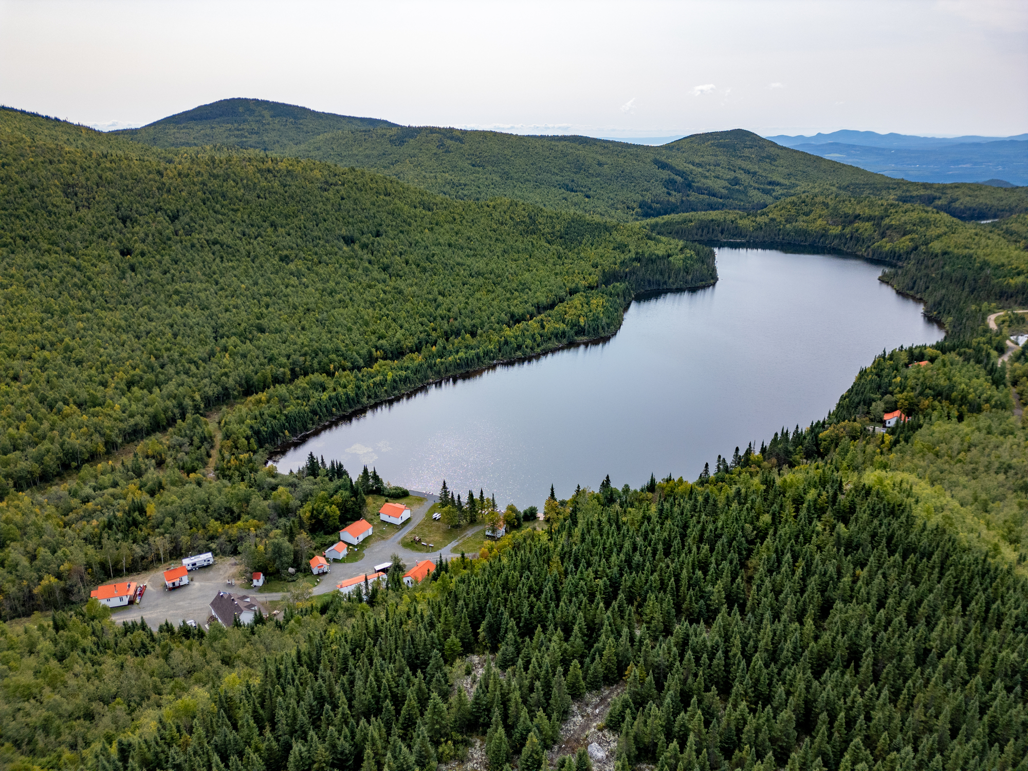 Aerial view of a serene lake surrounded by lush green forests and cozy cabins at Club des Trois Castors in Quebec