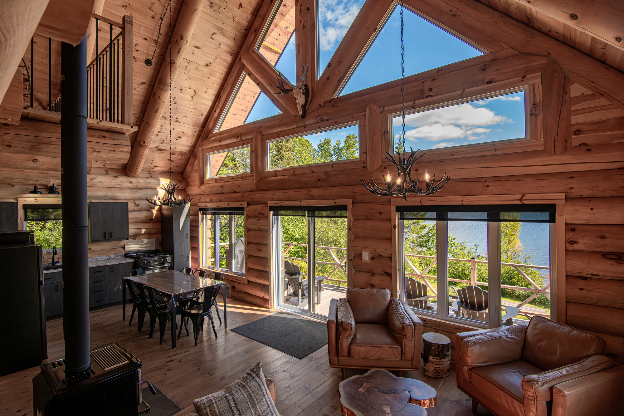 Cozy wooden cabin interior with large windows overlooking a lake, featuring comfortable seating and a dining area