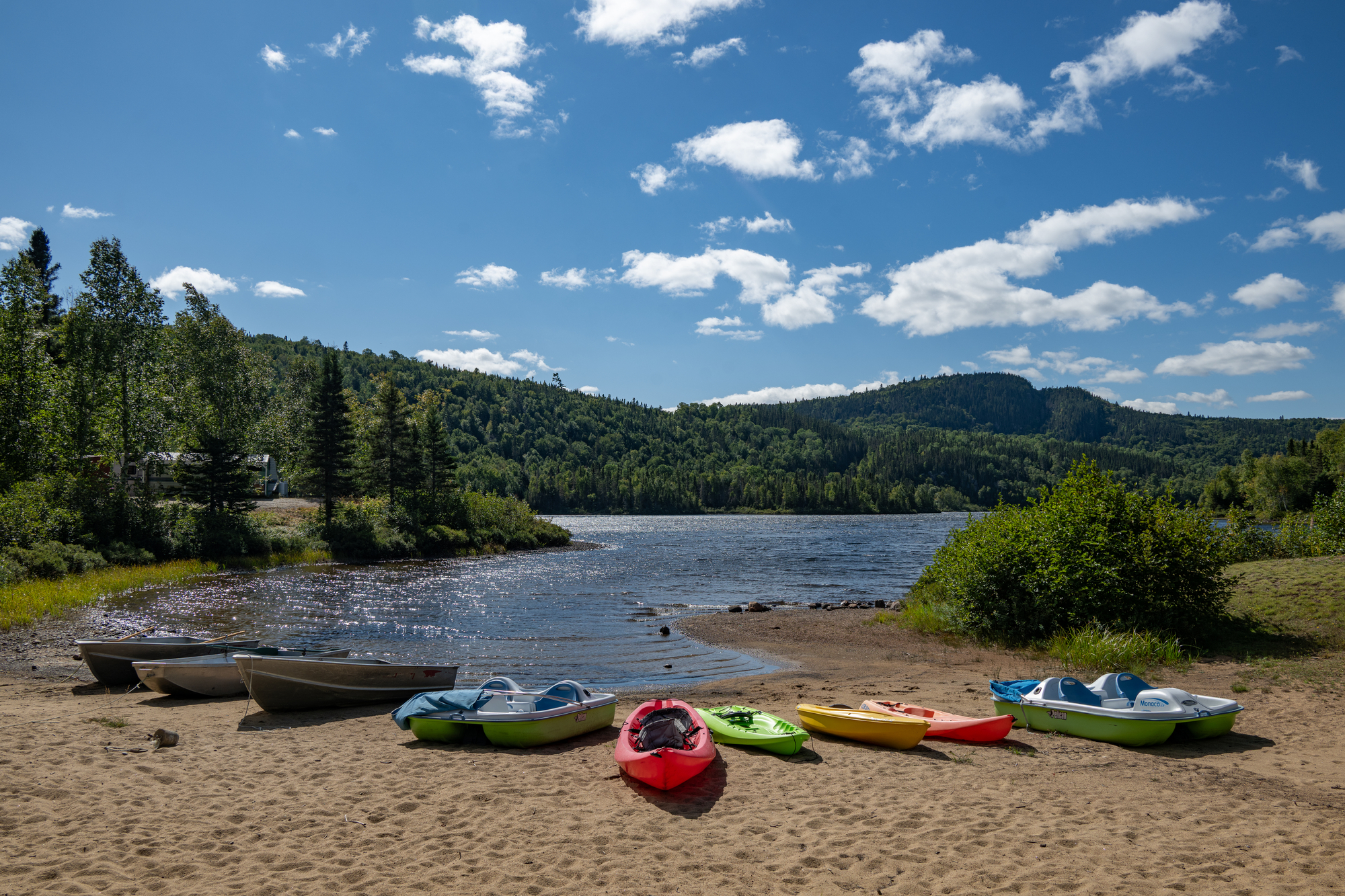 Kayaks colorés et canoës alignés sur la plage de sable, avec un lac et des collines verdoyantes en arrière-plan
