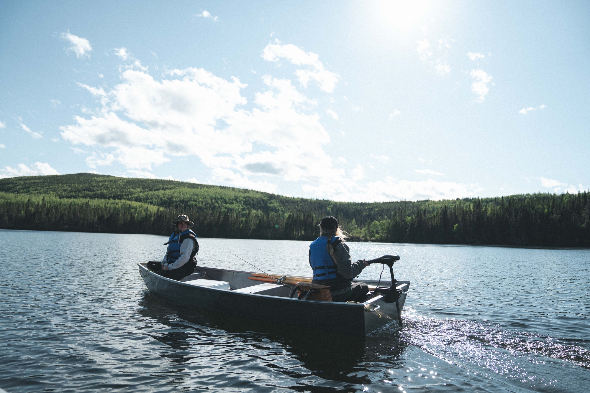 two people in a boat fishing on a calm lake surrounded by lush green forests and a clear blue sky