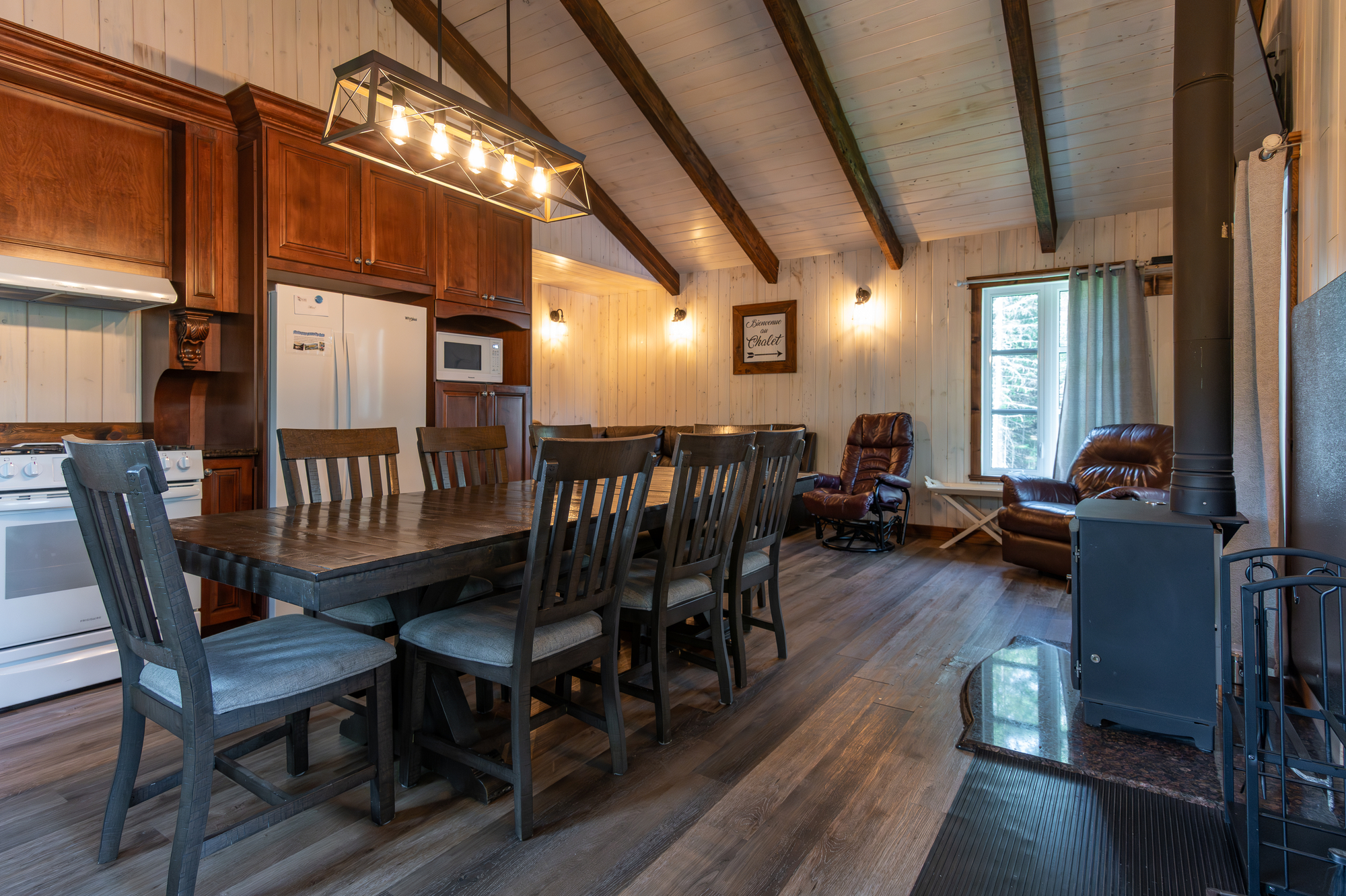 Cuisine spacieuse avec table en bois pour dix personnes, fauteuils en cuir, fenêtres lumineuses et poêle à bois dans chalet de vacances