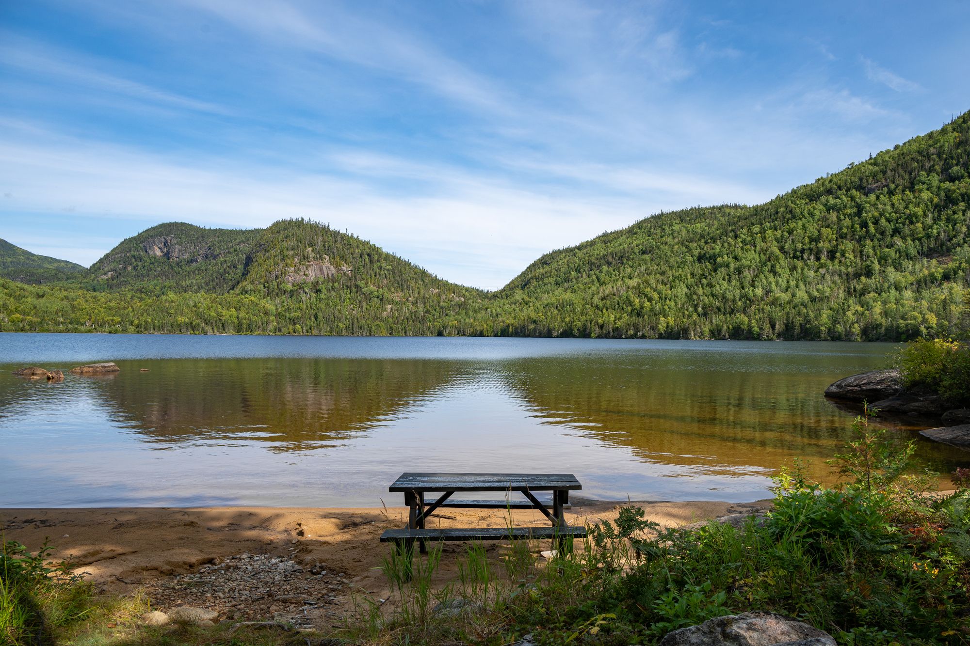Serene lakeside view with a picnic table, surrounded by lush green mountains and calm water in Charlevoix