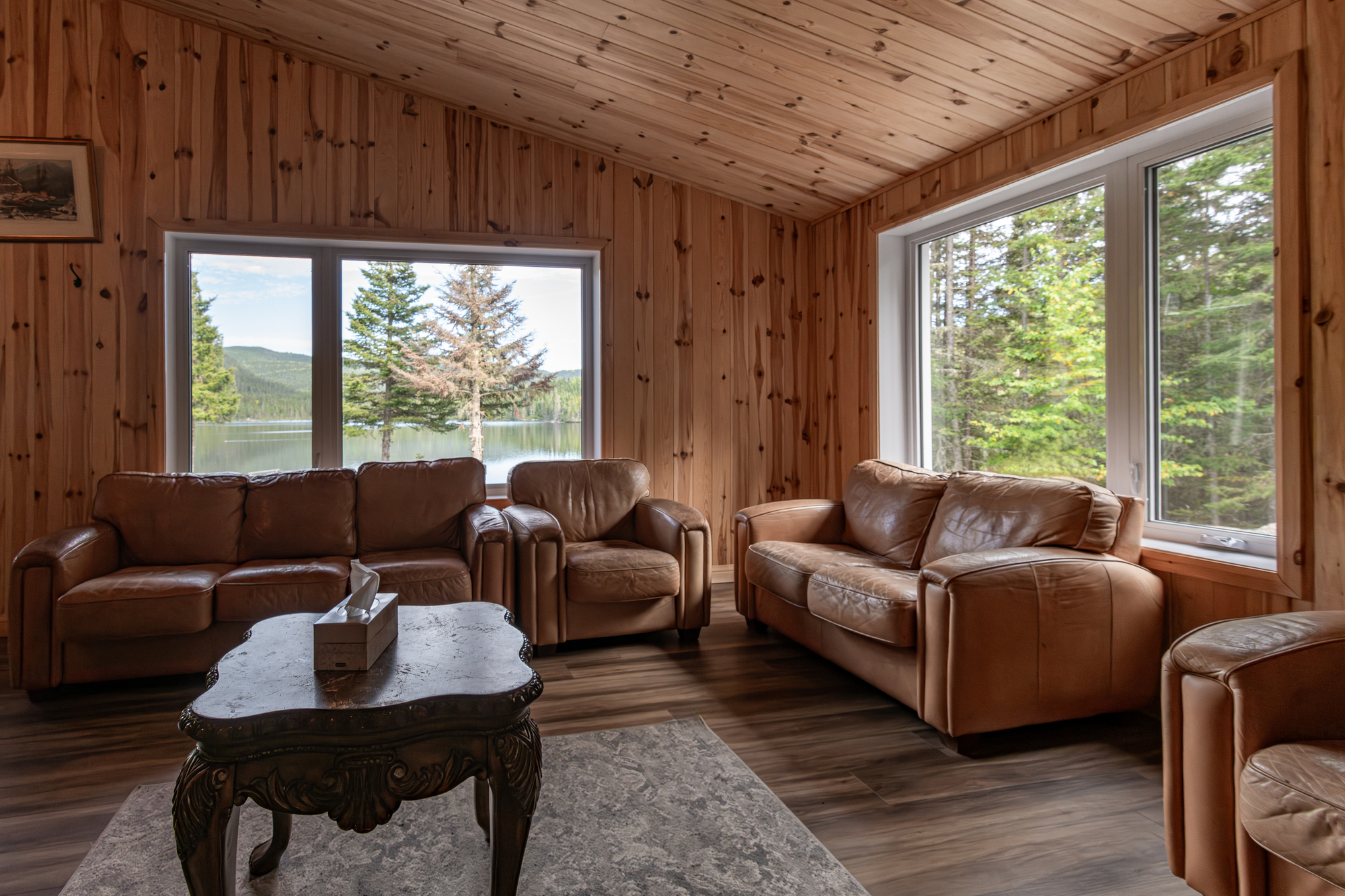 Cozy living area with leather sofas, wooden walls, and large windows overlooking a serene lake and forest in Charlevoix