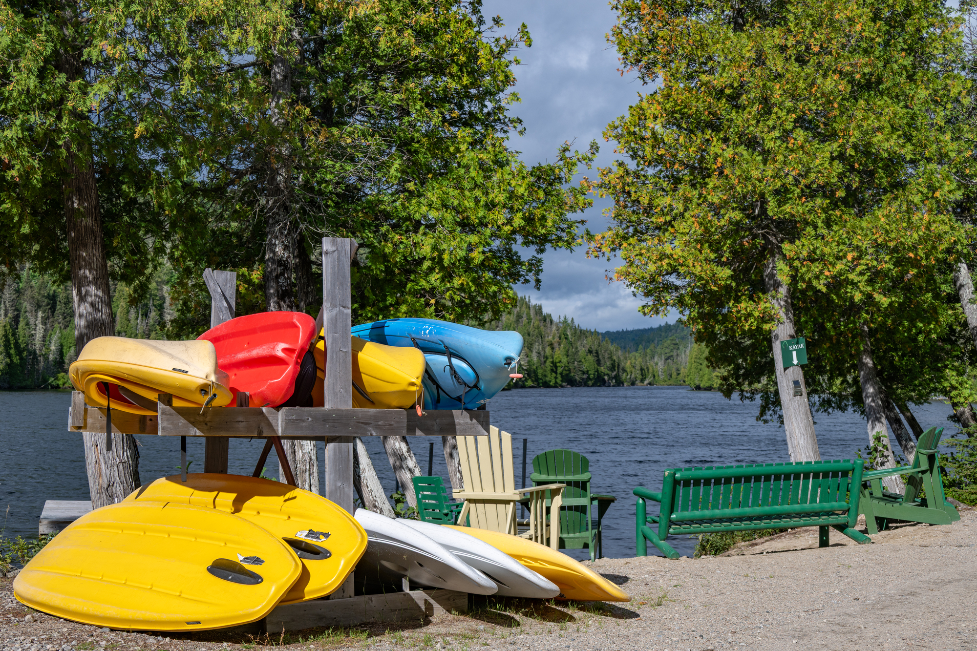 Kayaks colorés empilés sur un support près d'un lac entouré d'arbres, chaises vertes visibles pour se reposer