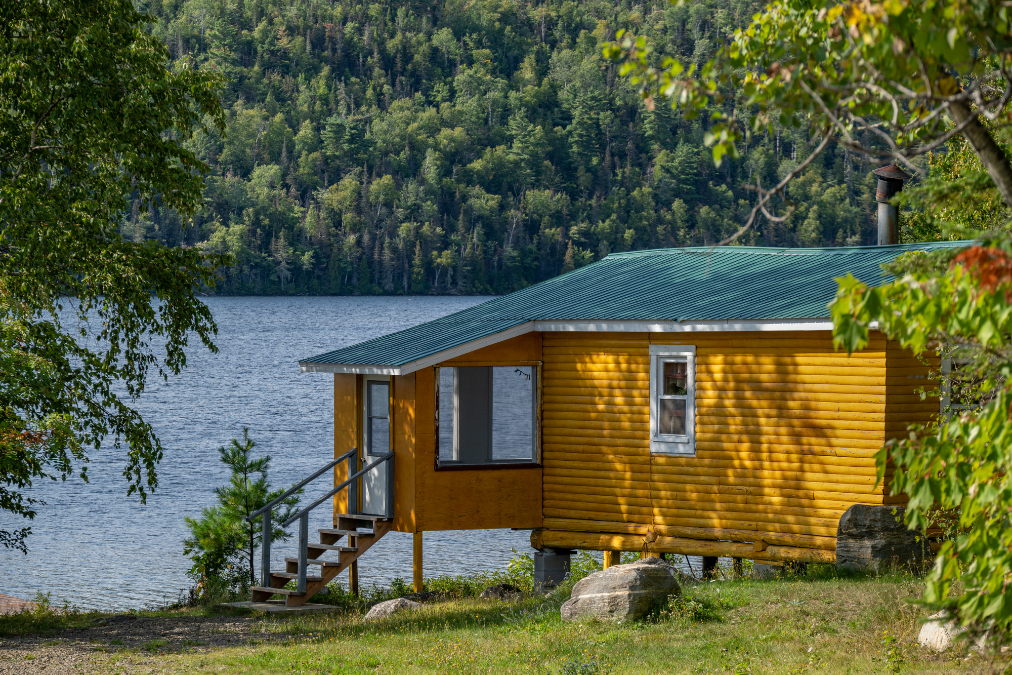 Yellow waterfront cabin with a green roof beside a serene lake surrounded by lush trees in Charlevoix Quebec