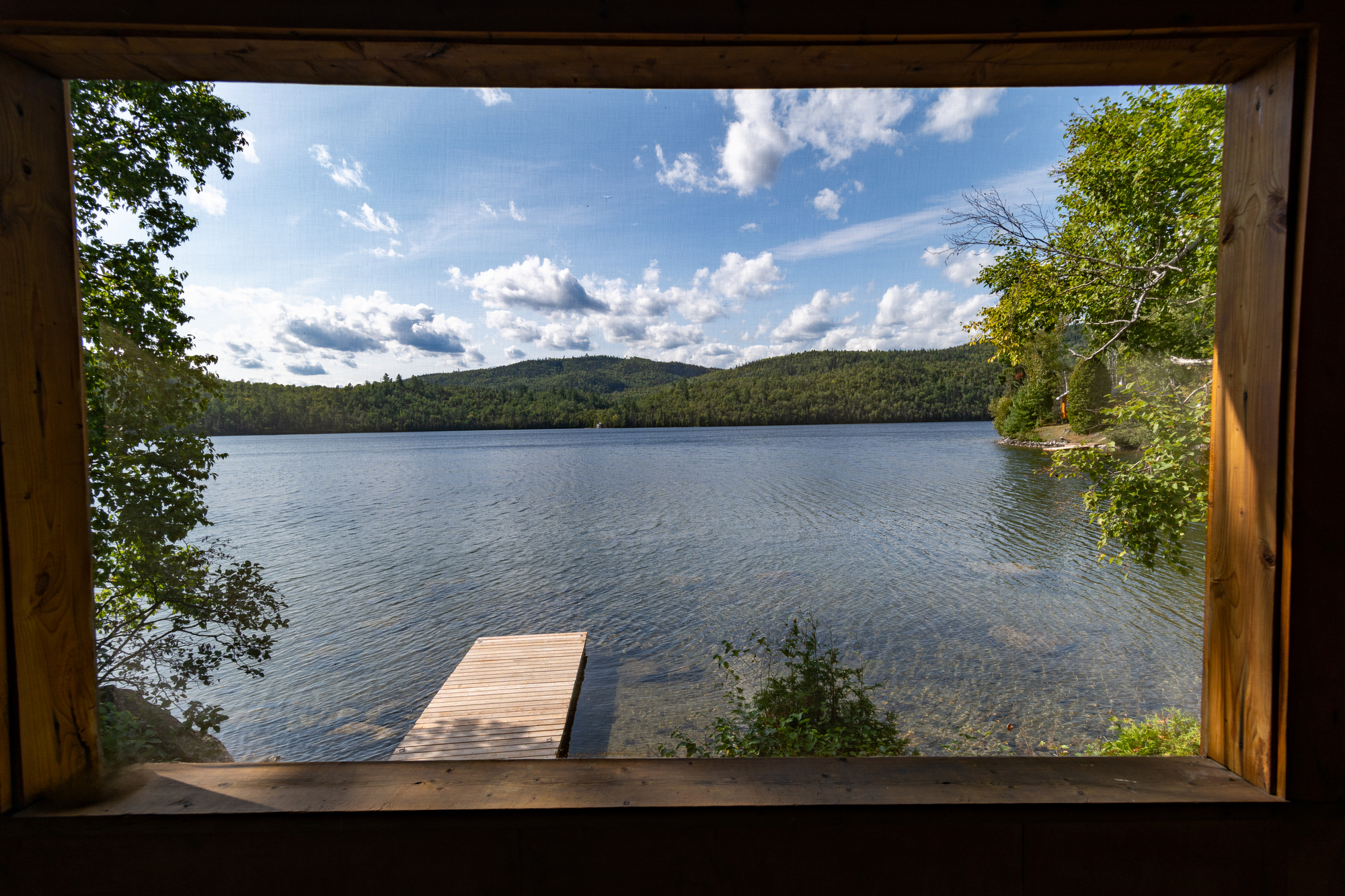 view of a serene lake framed by rustic wooden structure, surrounded by lush greenery and distant hills in Charlevoix