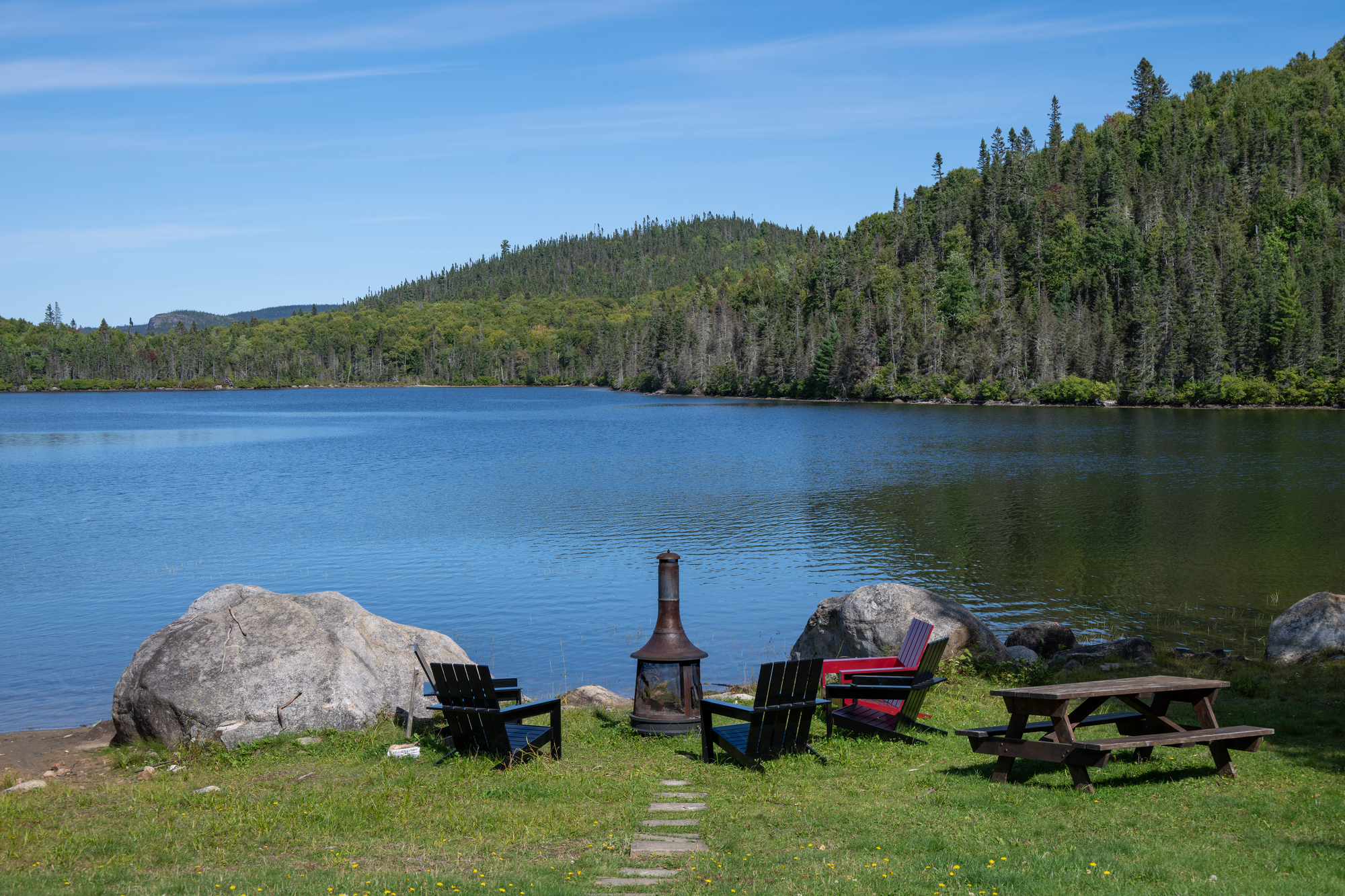 Lakeside setting with chairs around a fire pit, surrounded by trees and mountains under a clear blue sky