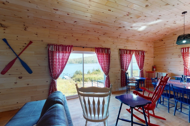 Interior of a cozy cottage with wooden walls, large windows overlooking a lake, colorful chairs, and a dining table