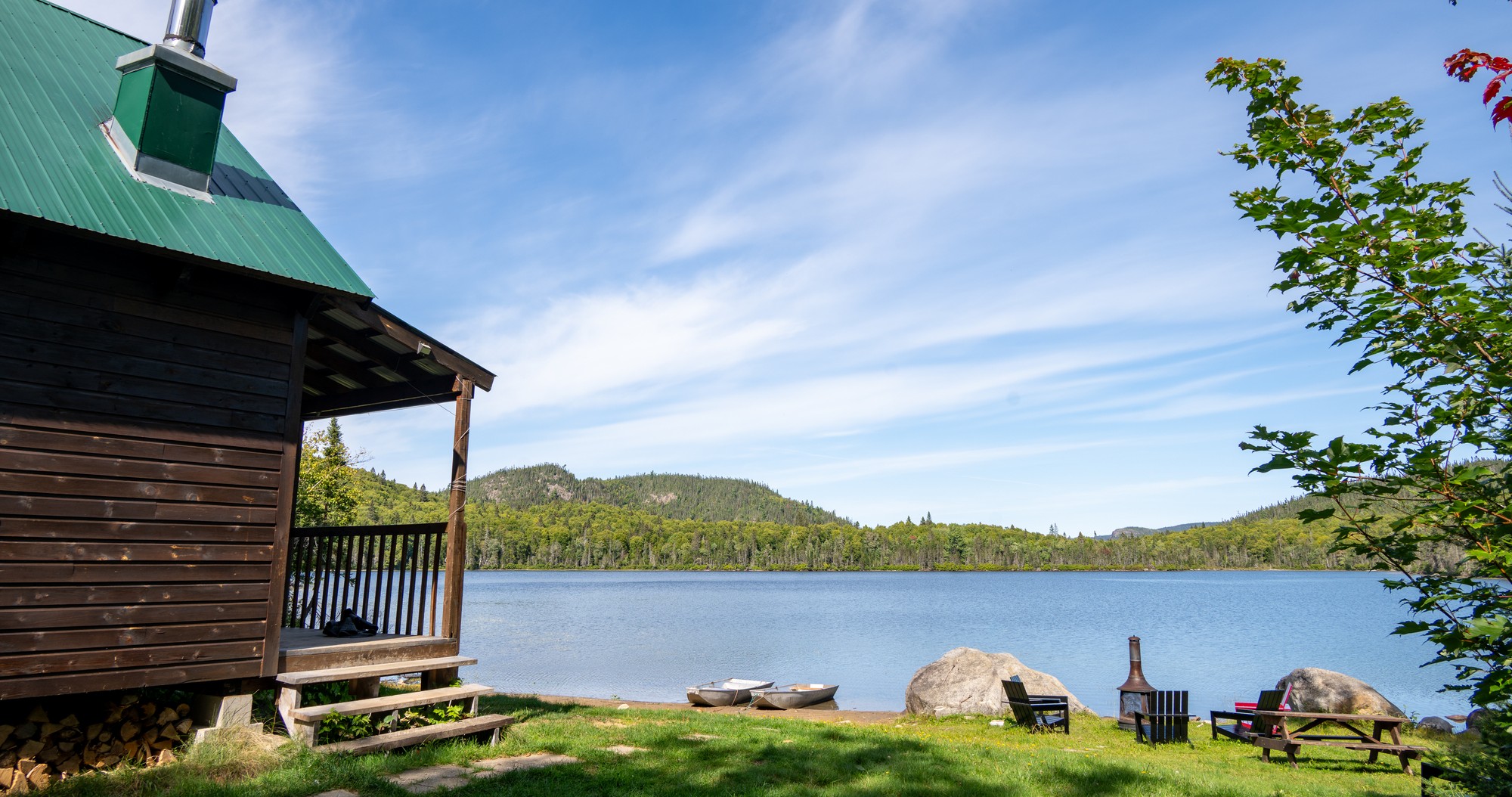 Cabane en bois au bord d'un lac, entourée d'arbres et de rochers, avec chaises sur la pelouse pour se détendre au soleil