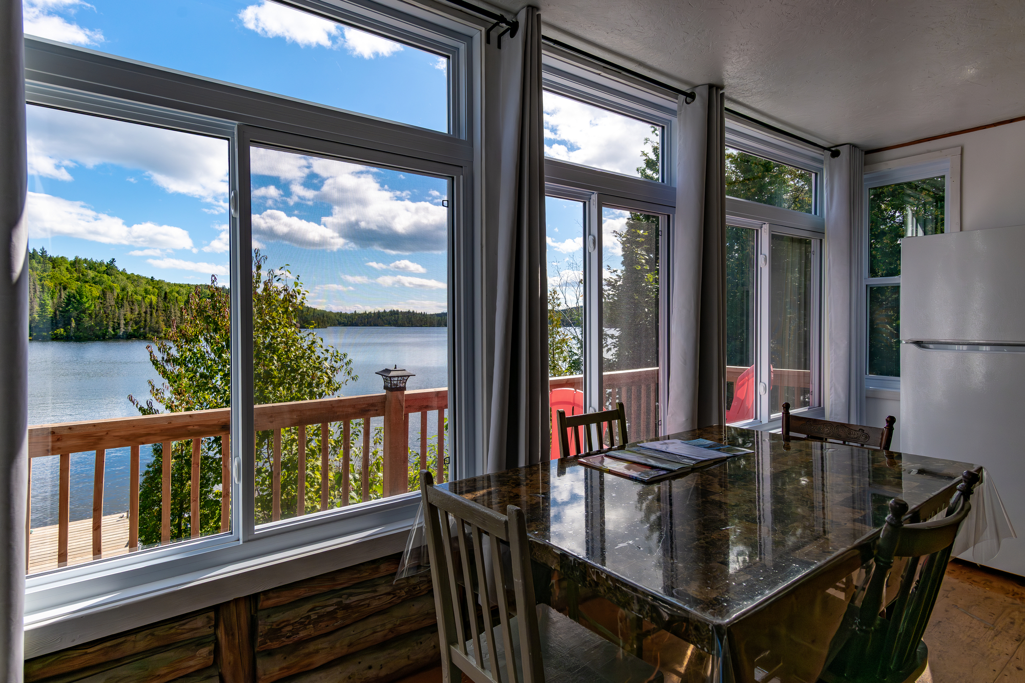 Vue intérieure d'un chalet avec une table pour manger, grandes fenêtres donnant sur un lac et la forêt environnante