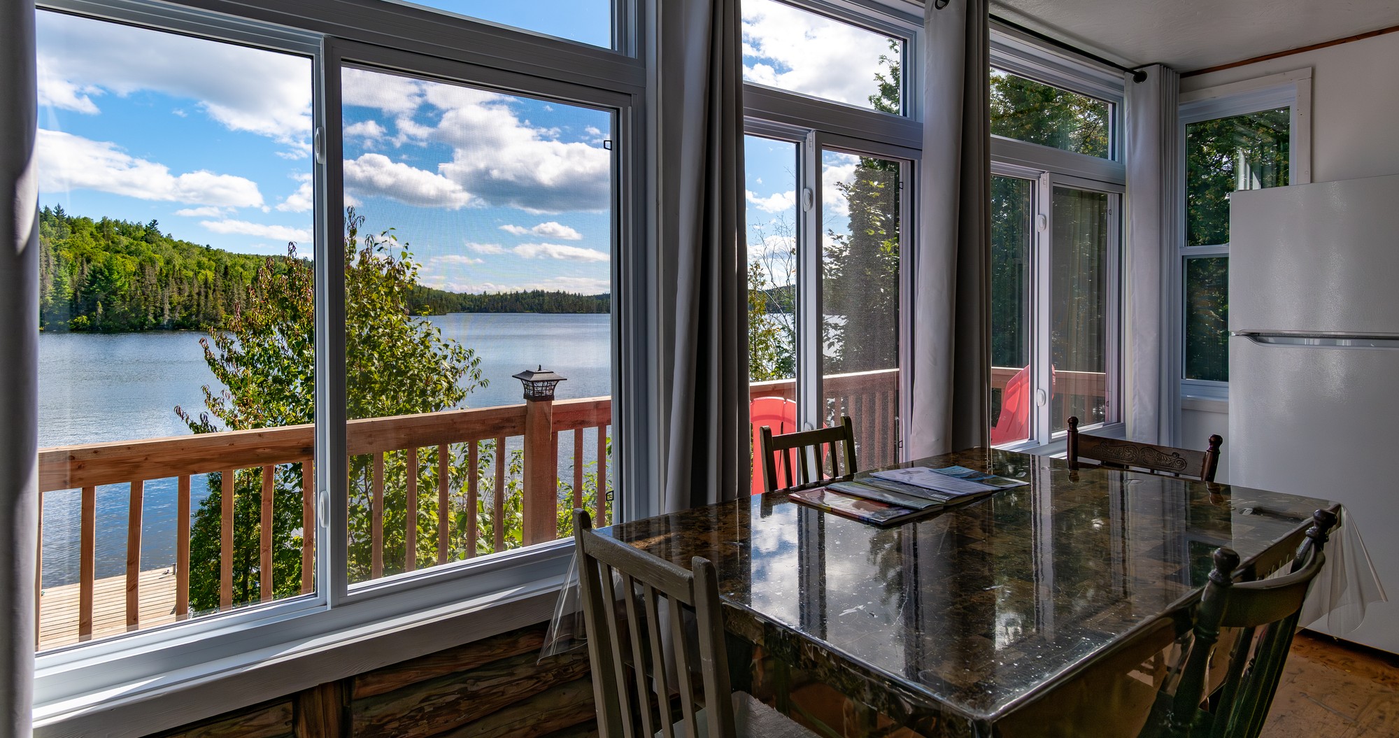 Vue d'un lac à Baie-Sainte-Catherine depuis une salle à manger lumineuse avec une table en bois et de grandes fenêtres