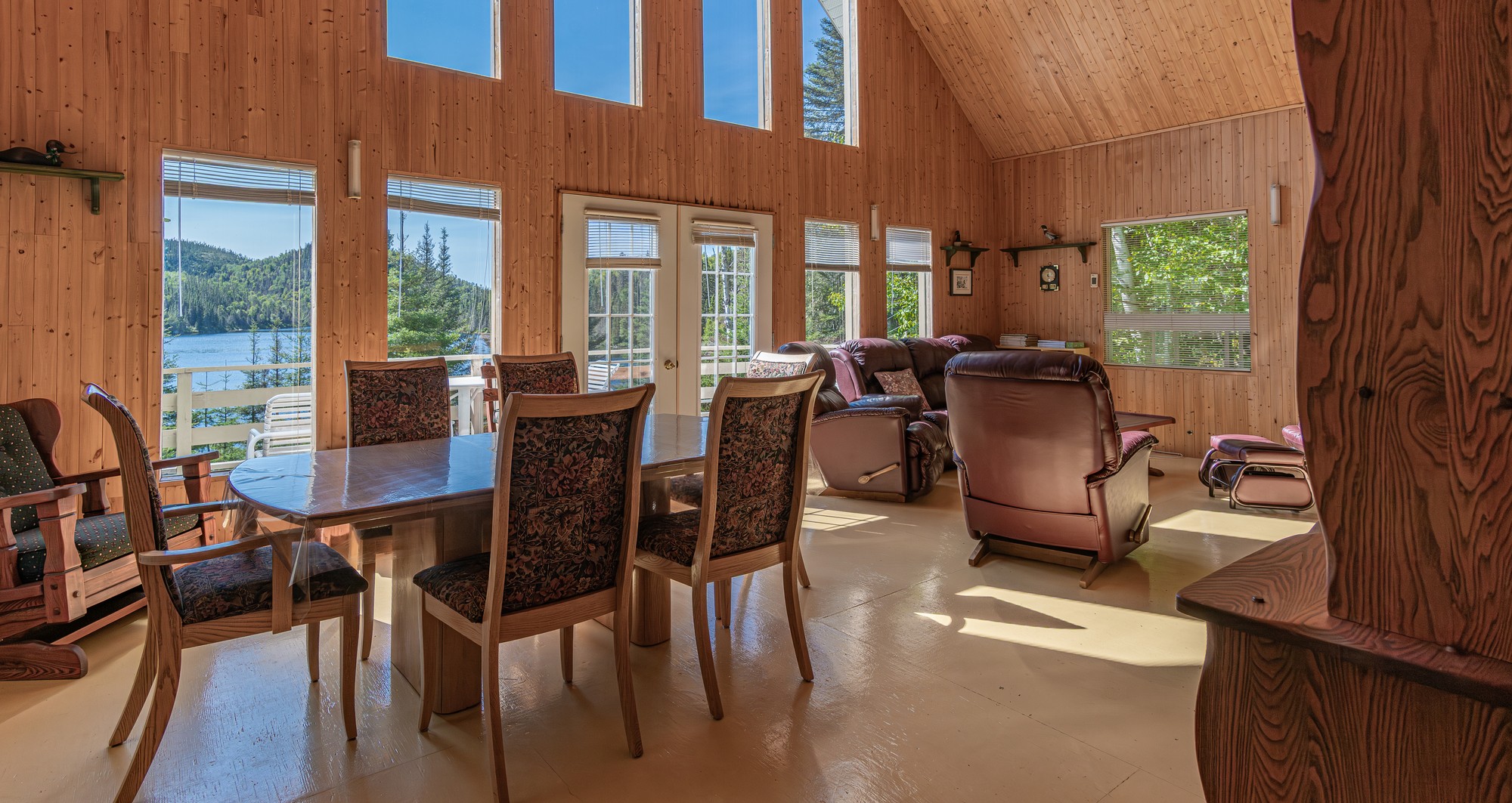 Intérieur lumineux d'un chalet avec tables et chaises en bois, fauteuils confortables et vue sur le lac et la nature environnante