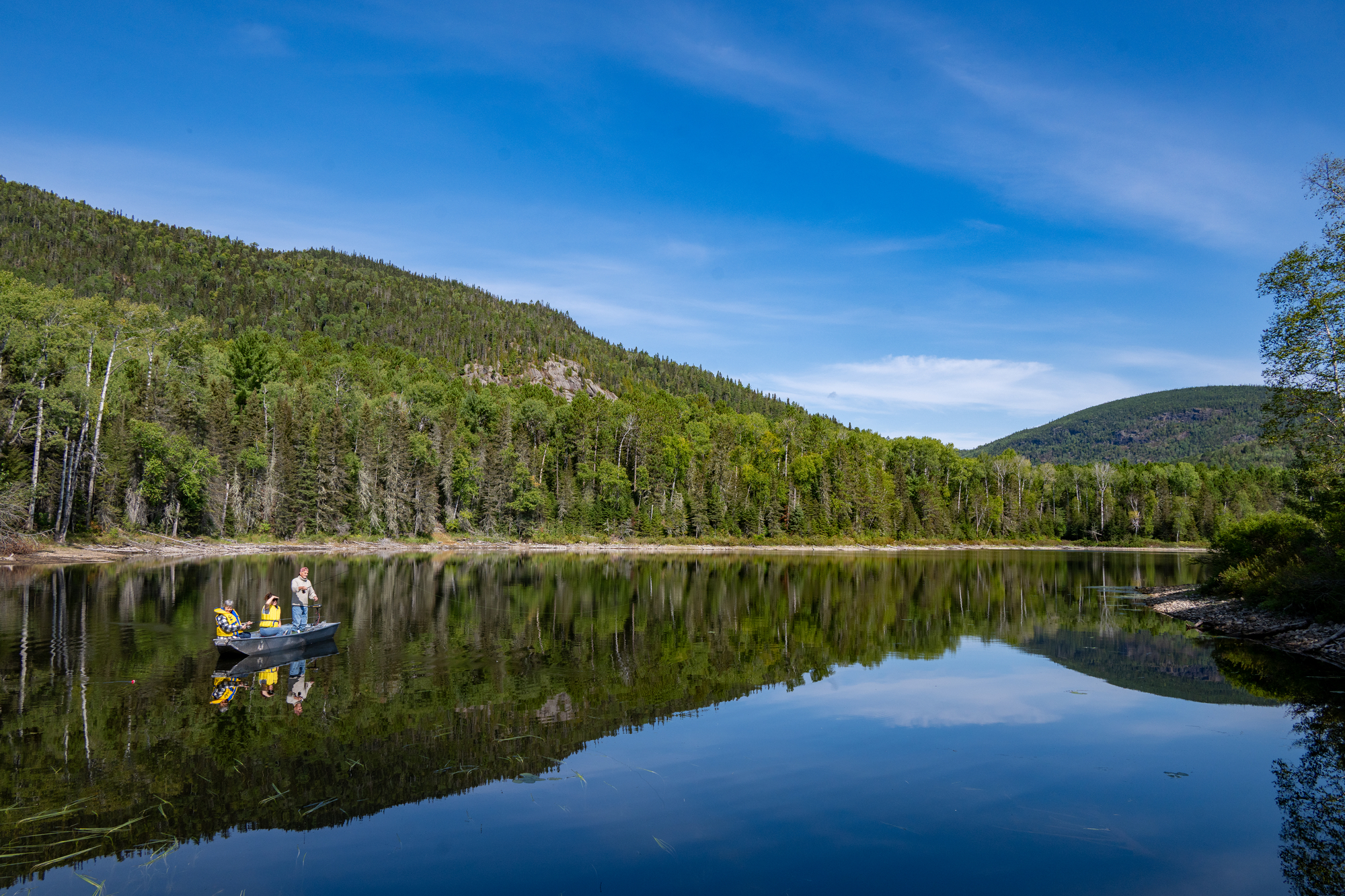 Deux personnes pêchent sur un lac calme entouré de forêts verdoyantes sous un ciel bleu clair